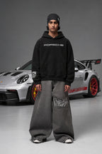 Full front view of the black Porsche hoodie beside a Porsche car